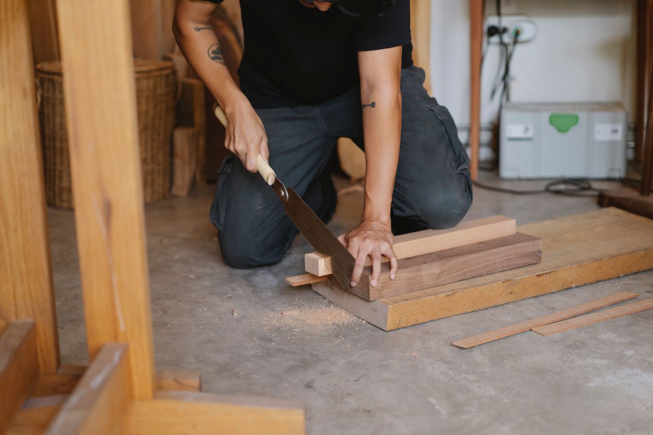 about-us Unrecognizable skilled male carpenter with tattoo cutting wooden planks with hand saw while sitting on floor in professional studio during work